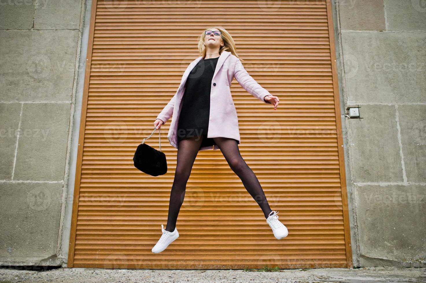 loira de óculos e casaco rosa com bolsa saltar contra persianas. foto