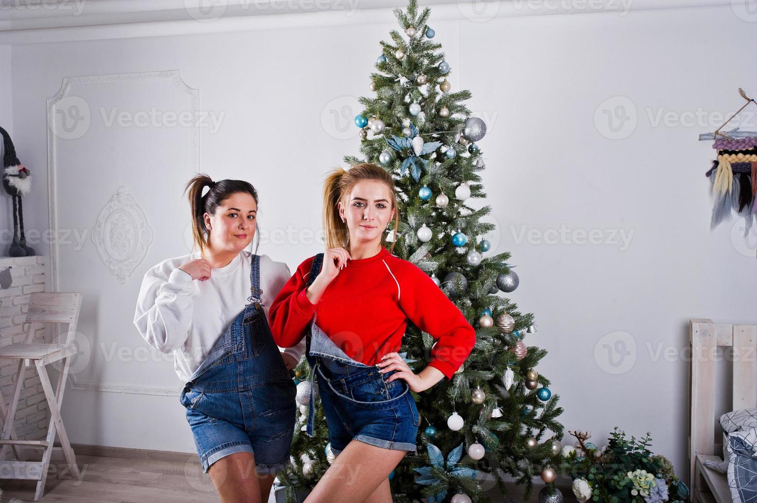 duas amigas bonitas e divertidas usam shorts jeans de macacão e polainas contra árvore de ano novo com decoração de chrisrmas. foto