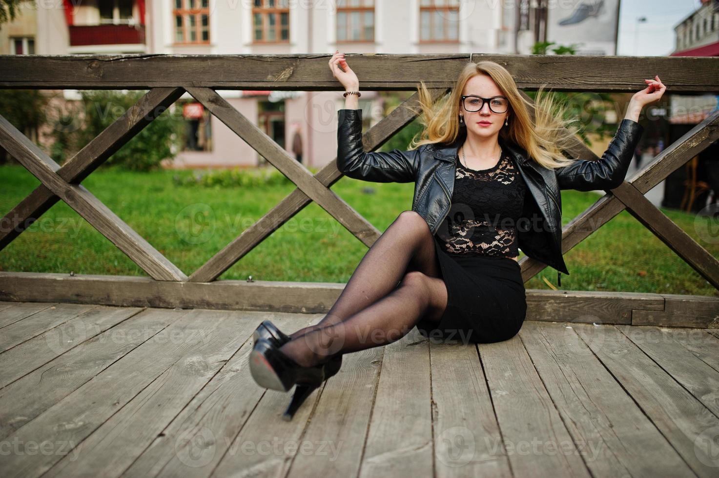 menina loira elegante usar jaqueta de couro preta posando nas ruas da cidade, sentado na madeira. foto