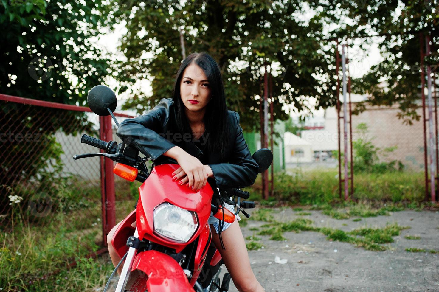 retrato de uma mulher legal e incrível de vestido e jaqueta de couro preta, sentado em uma moto vermelha legal. foto