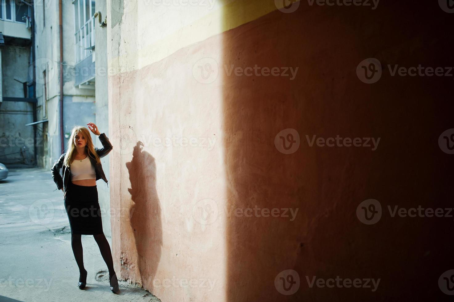 menina loira elegante usar jaqueta de couro preta posando nas ruas da velha parede de fundo da cidade com sombras. foto