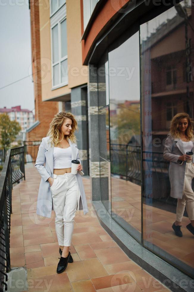 menina elegante modelo loira encaracolada usar branco com uma xícara de café na mão posando contra a grande janela. foto