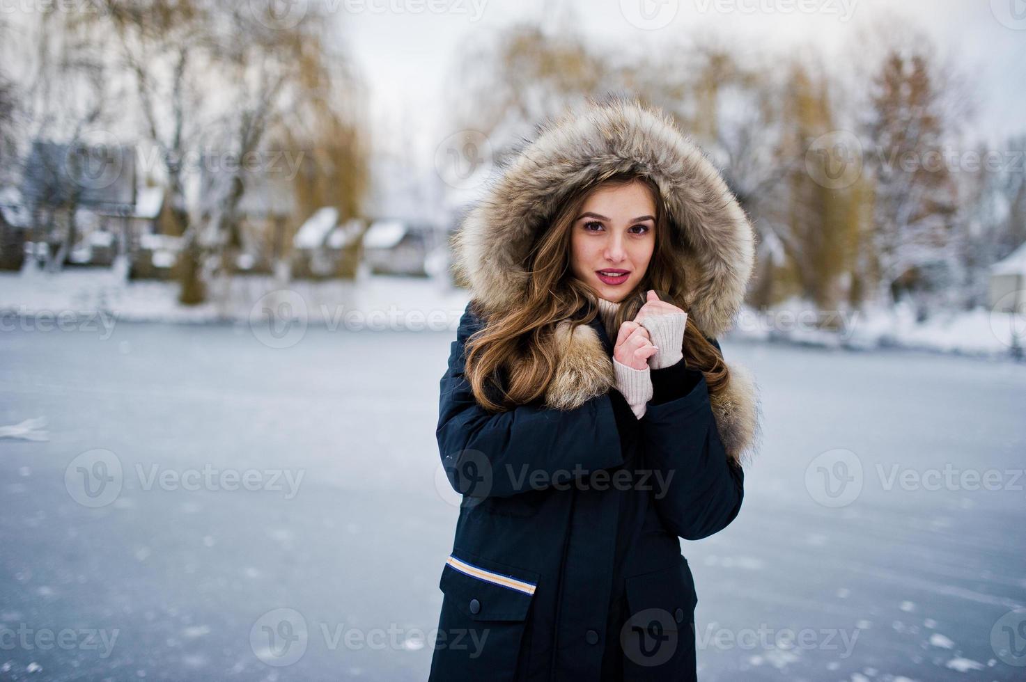 linda menina morena em roupas quentes de inverno. modelo na jaqueta de inverno contra o lago congelado no parque. foto