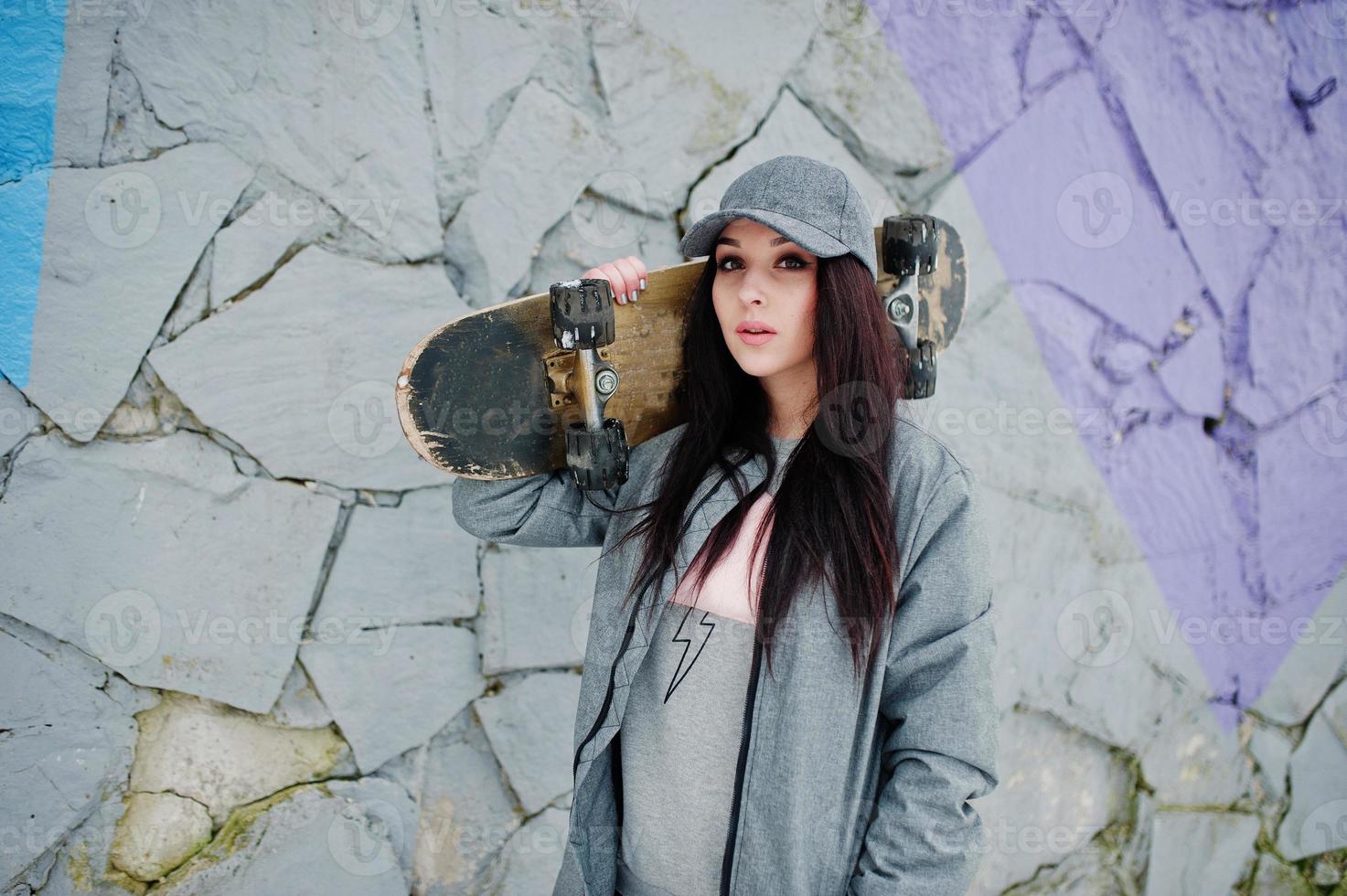 menina morena elegante boné cinza, estilo casual de rua com skate em dia de inverno contra parede colorida. foto