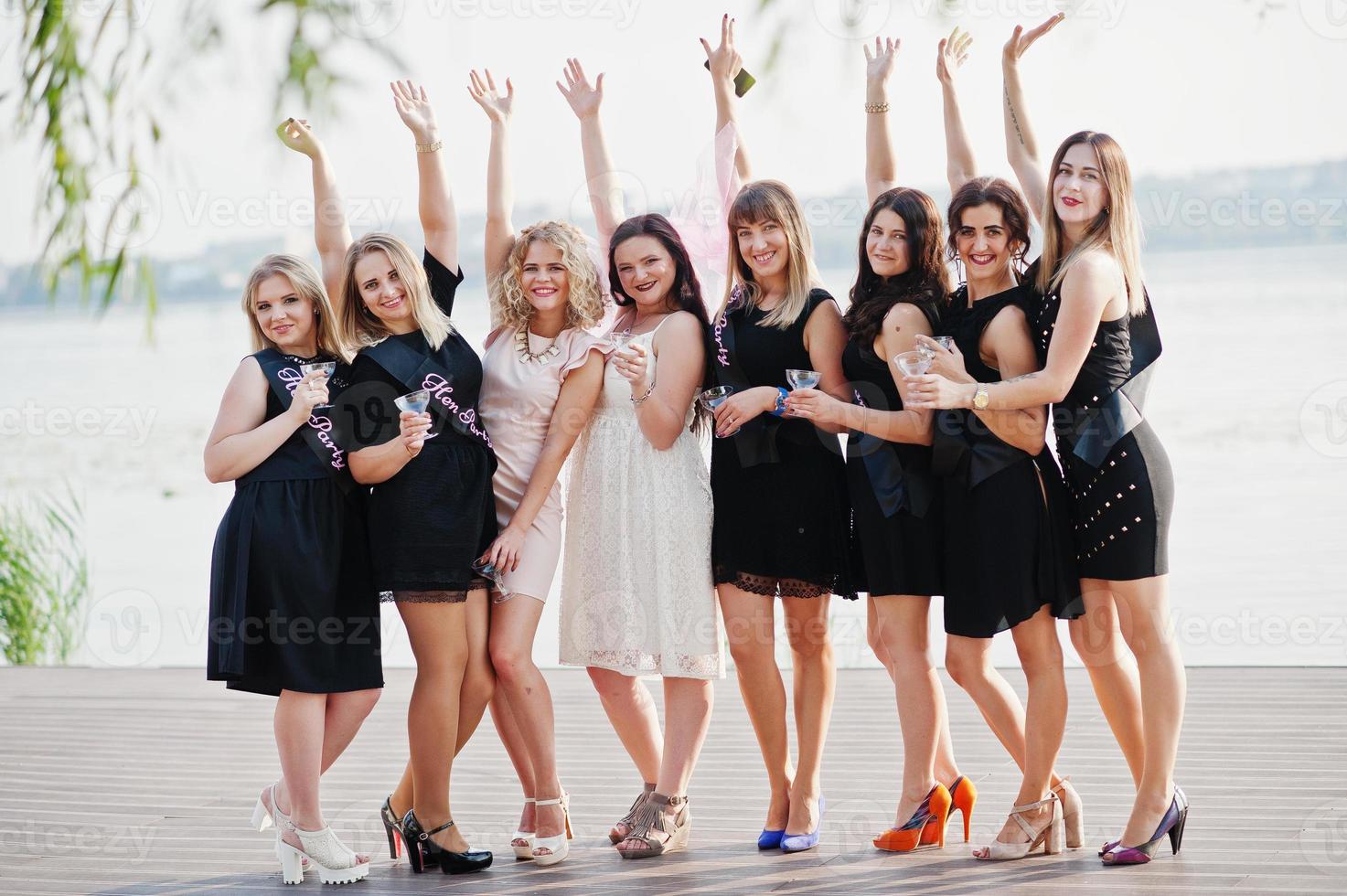 grupo de 8 meninas usam preto e 2 noivas na festa de despedida contra praia ensolarada bebendo champanhe. foto