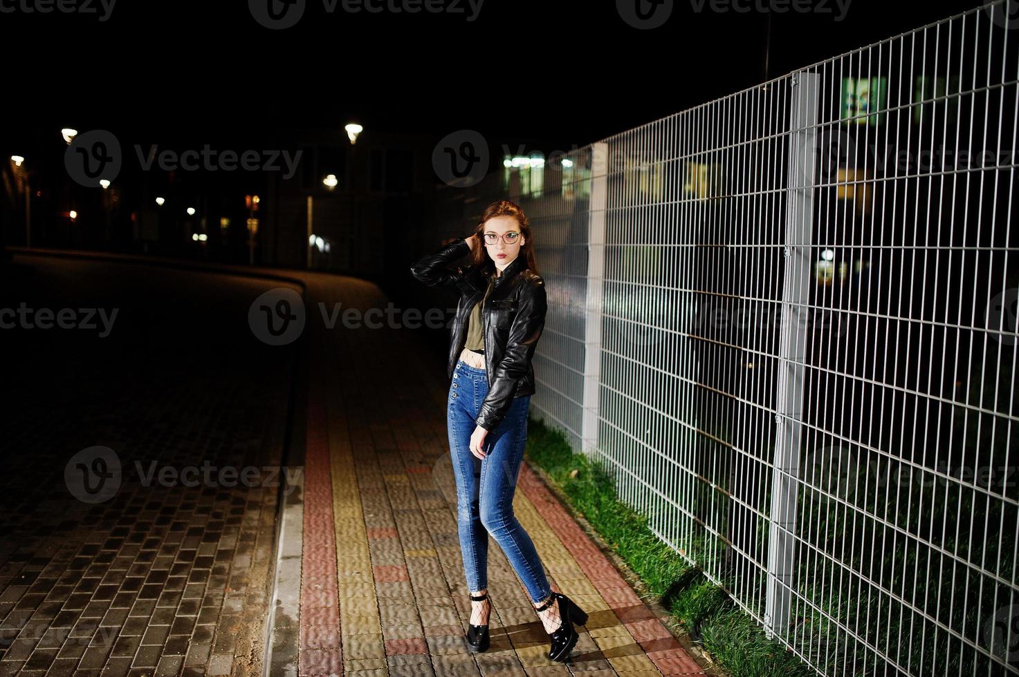 retrato noturno do desgaste do modelo de menina em jeans e jaqueta de couro contra cerca de ferro. foto