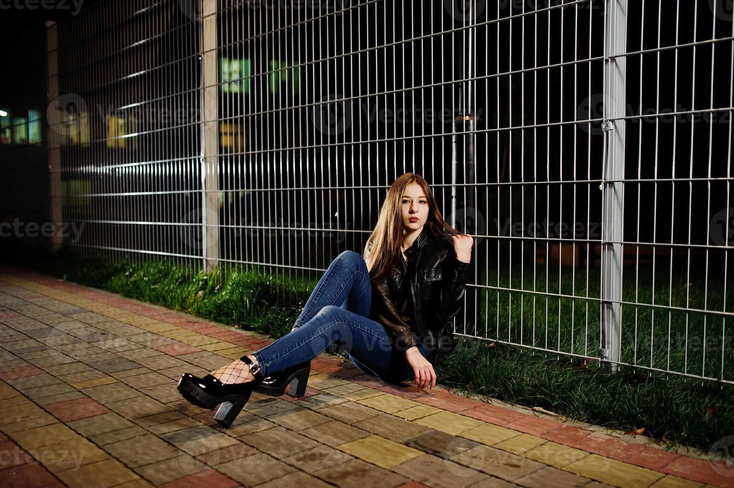 retrato noturno do desgaste do modelo de menina em jeans e jaqueta de couro contra cerca de ferro. foto