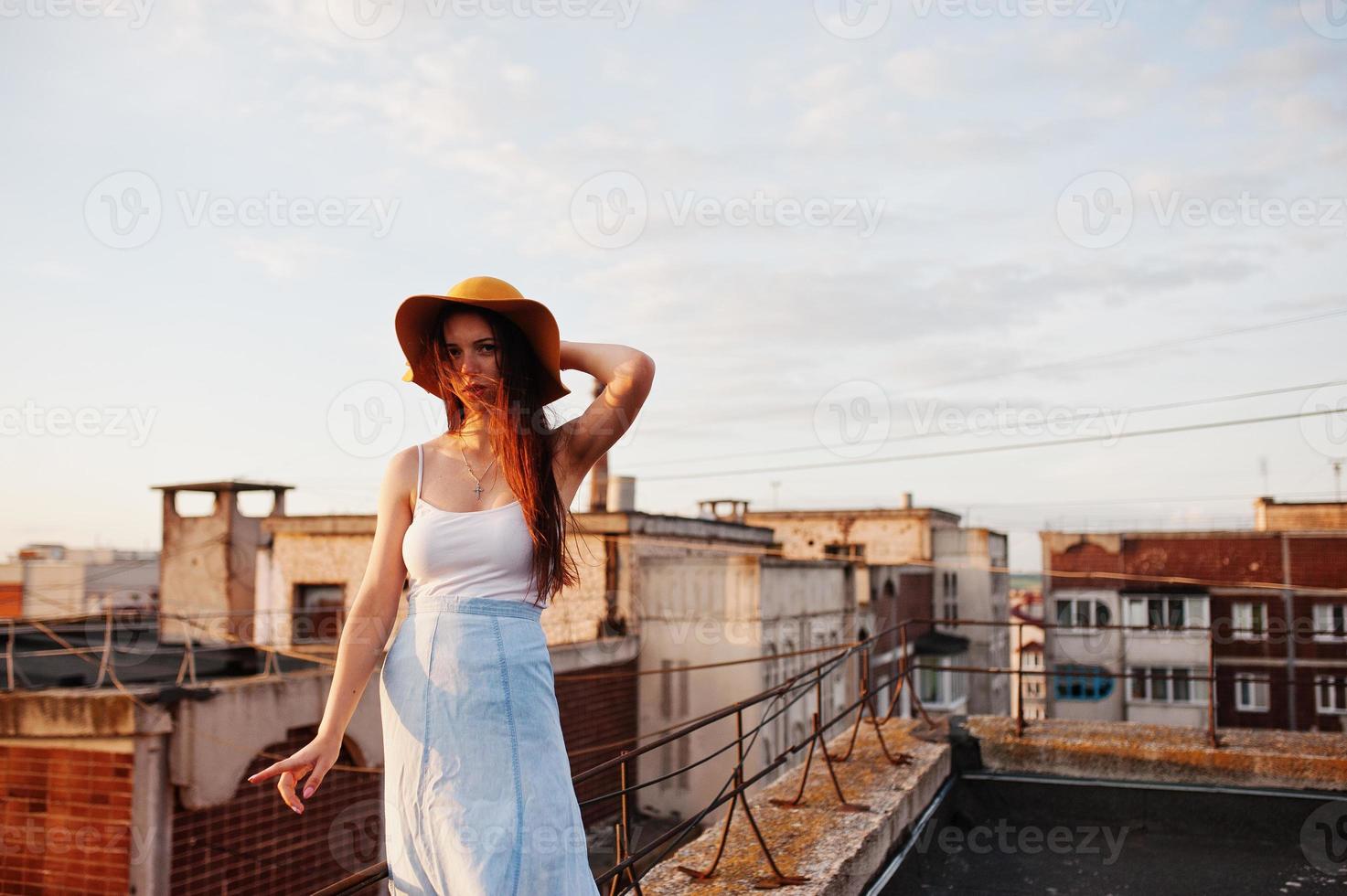 retrato de uma bela jovem de camiseta branca e saia azul posando no telhado com seu chapéu laranja ao pôr do sol. foto