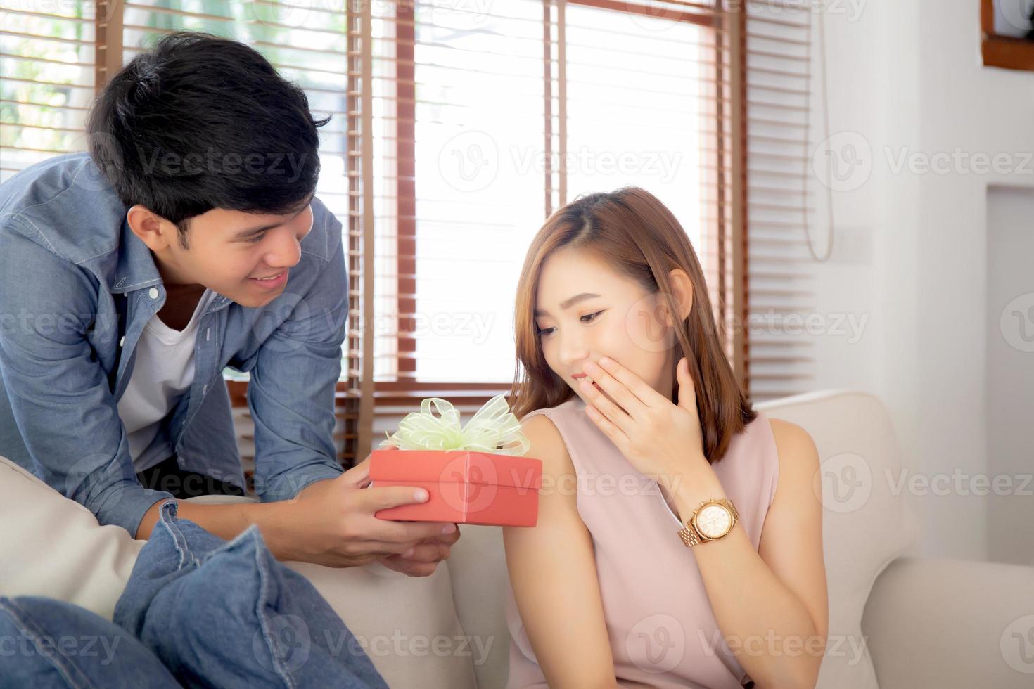 jovem casal asiático comemora aniversário juntos, homem asiático dando presente de caixa de presente para mulher para surpresa na sala de estar, feminino se sentindo feliz e animado aniversário, conceito de feriado dos namorados. foto