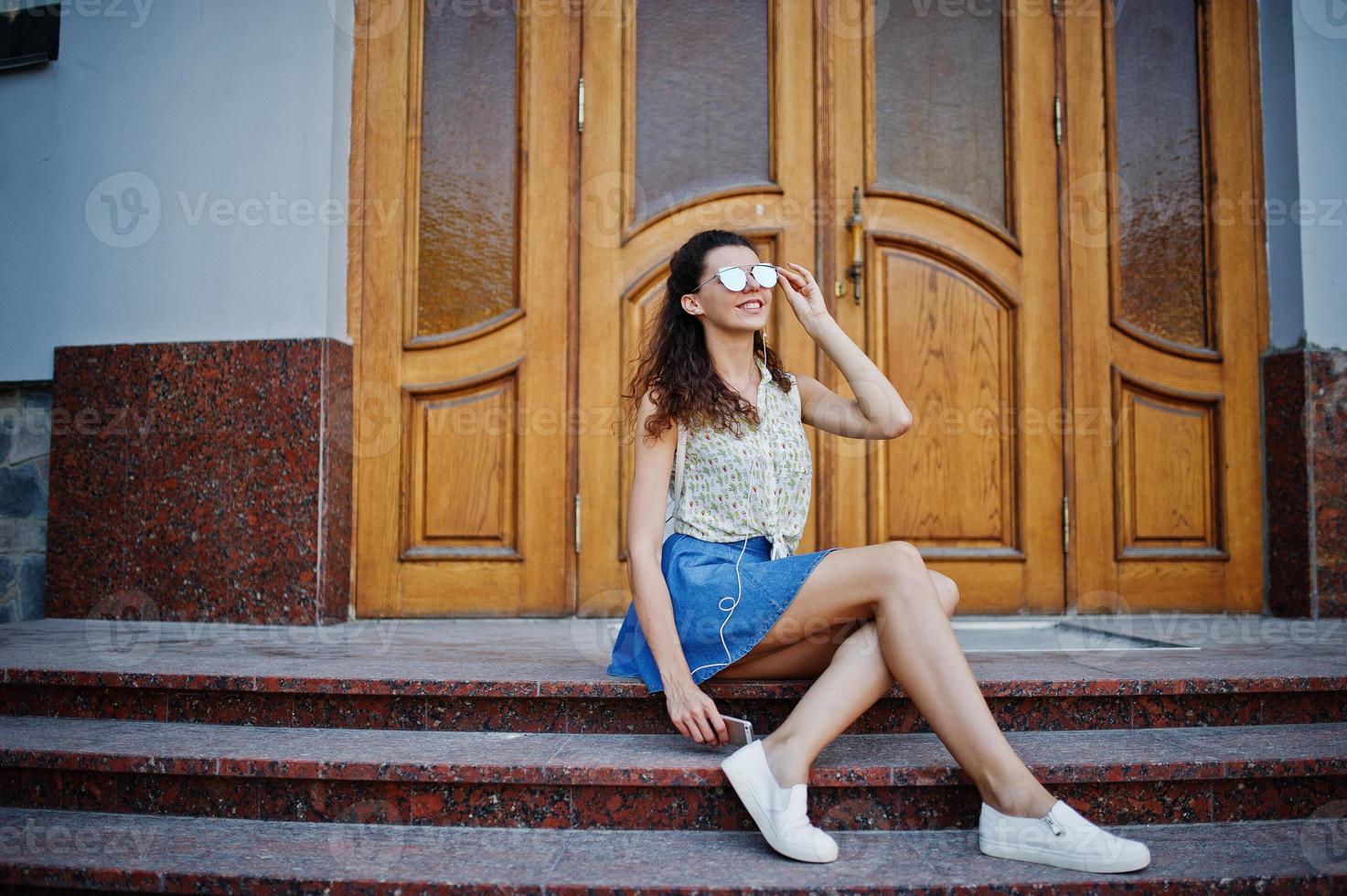 menina elegante encaracolada usa saia jeans azul, blusa e música de fones de ouvido. retrato nas ruas da cidade. foto