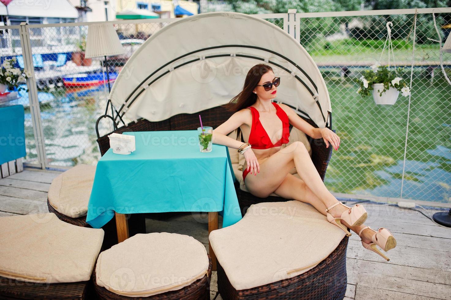 retrato de uma garota deslumbrante em maiô de biquíni vermelho e óculos de sol, aproveitando seu tempo sentado no sofá no cais à beira do lago. foto