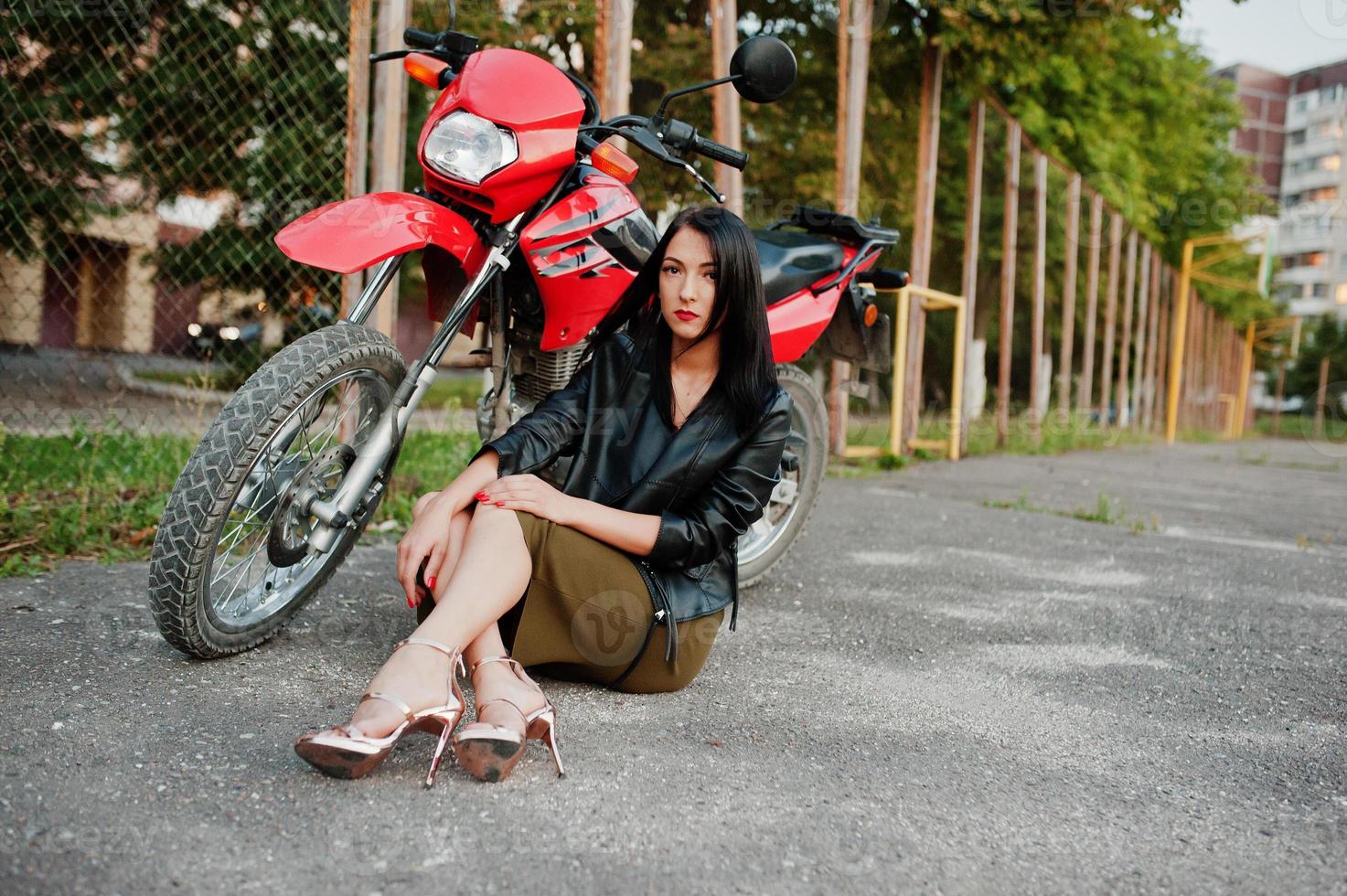 retrato de uma mulher legal e incrível de vestido e jaqueta de couro preta, sentado em uma moto vermelha legal. foto