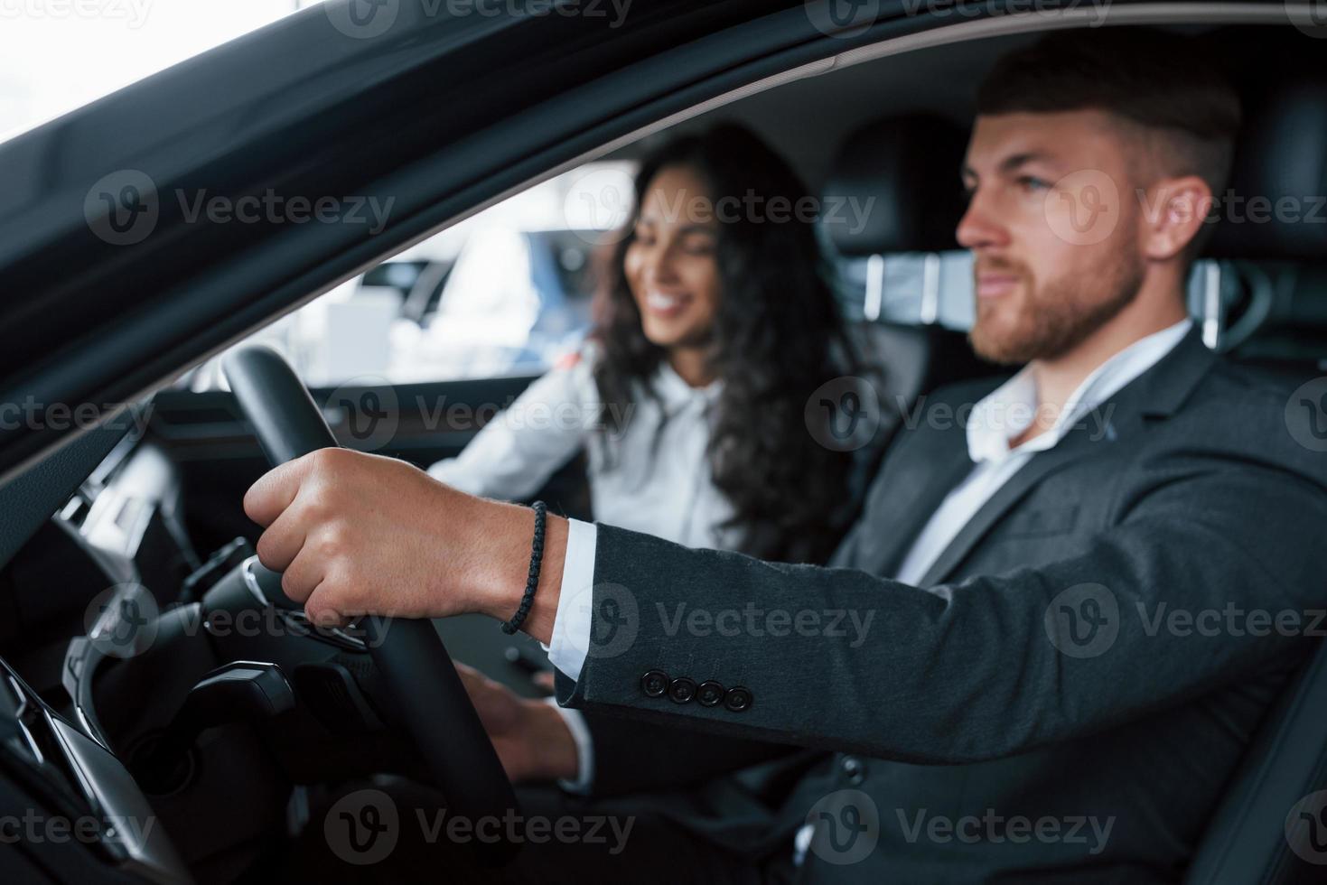 humor alegre. lindo casal bem sucedido tentando carro novo no salão de automóveis foto