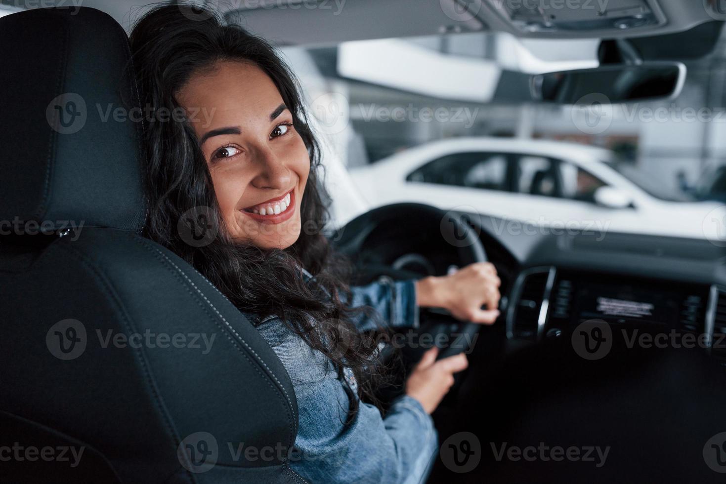 sorriso dentuço. linda garota com cabelo preto tentando seu novo carro caro no salão de automóveis foto