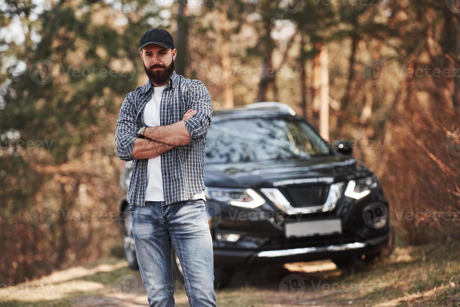 olhar confiante. homem barbudo perto de seu novo carro preto na floresta. conceito de férias foto