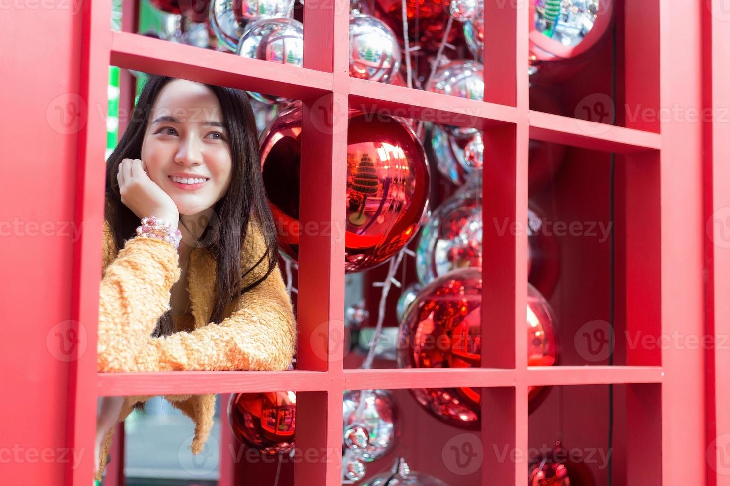 mulher bonita asiática de cabelos compridos vestindo um manto amarelo e sorriso feliz em pé na cabine telefônica vermelha no tema de celebrar o natal e feliz ano novo foto