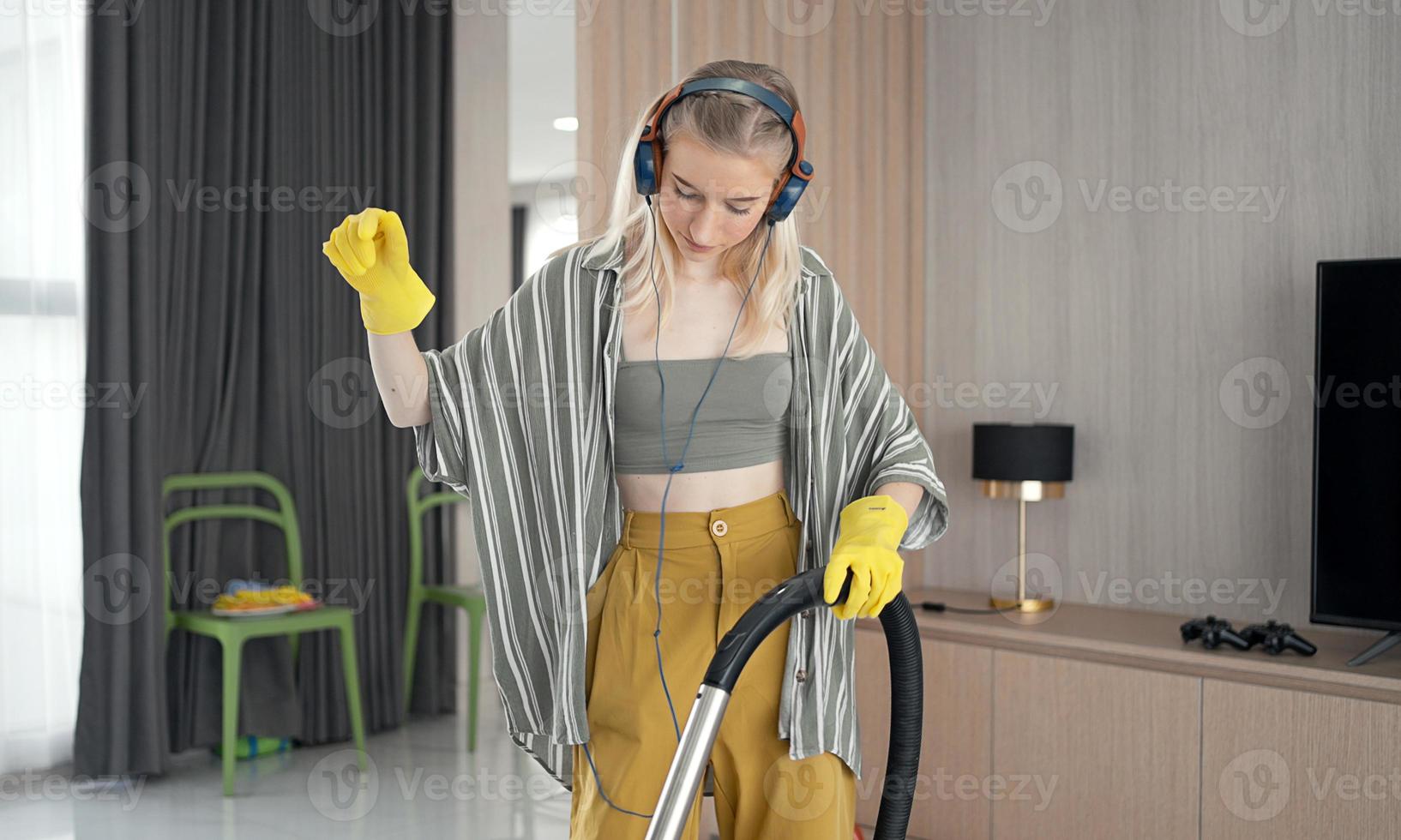 jovem se diverte enquanto limpa o chão com aspirador de pó. mulher feliz fazendo trabalhos domésticos em casa gosta de música usando fones de ouvido. foto