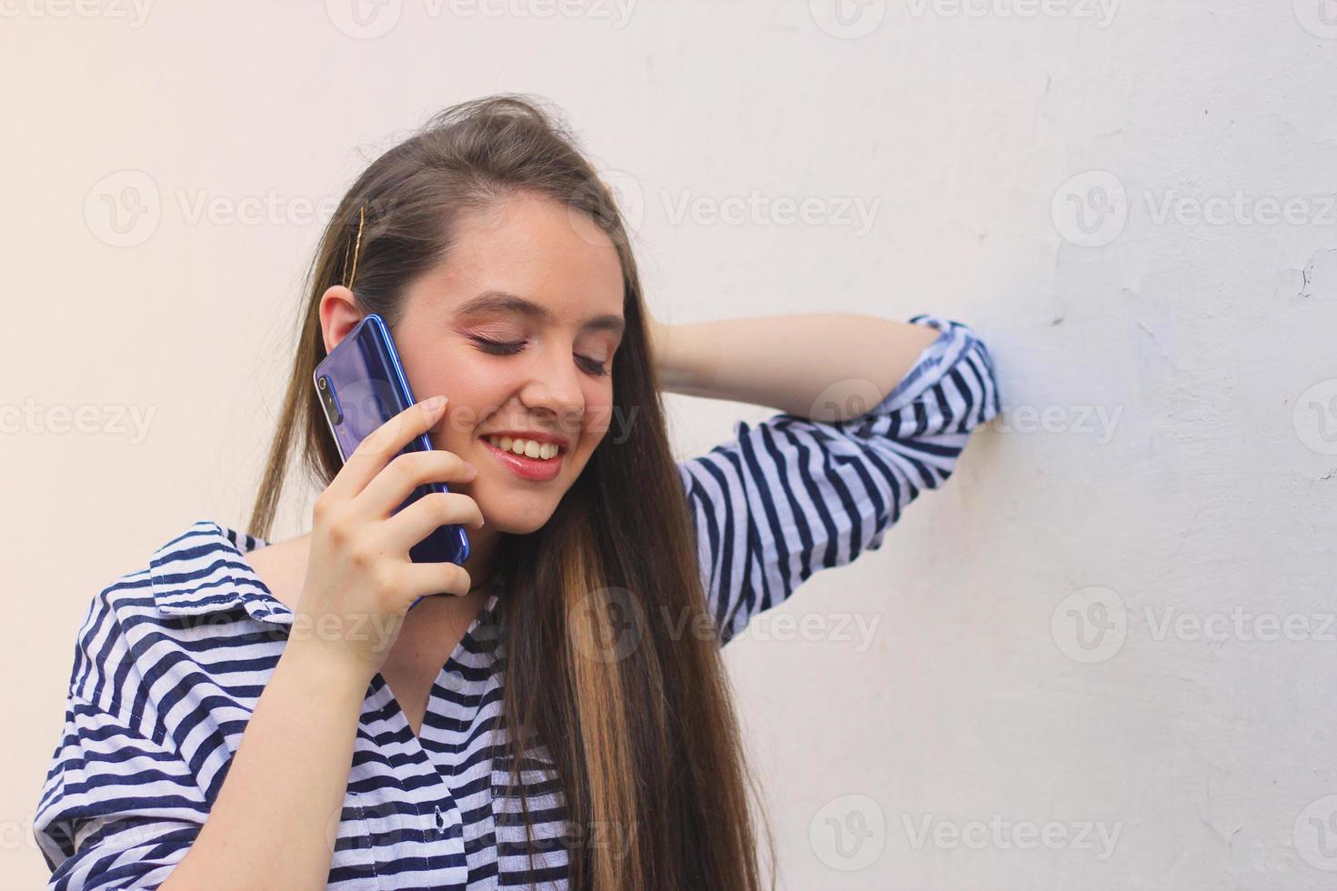 adolescente feliz falando ao telefone cem contra a parede, jovem com longos cabelos castanhos atende a chamada foto