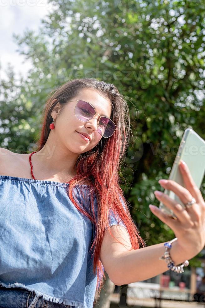 alegre mulher na moda com cabelo vermelho tomando café no parque, tomando selfie foto