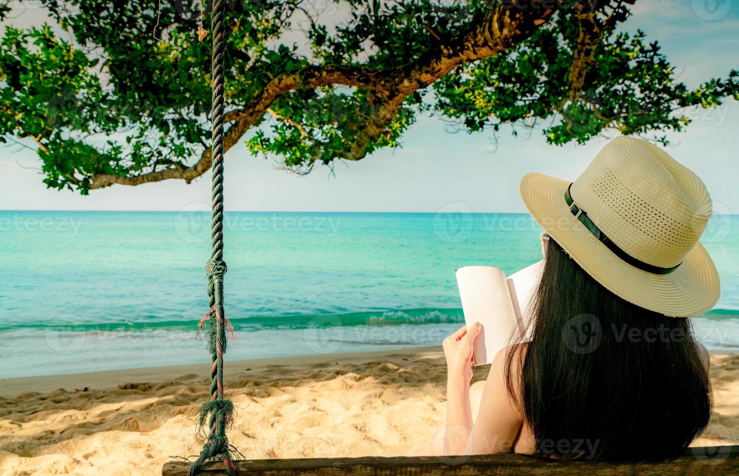 as mulheres sentam e lêem um livro em balanços debaixo da árvore à beira-mar. vista traseira da mulher asiática sexy com chapéu de palha relaxe e aproveite as férias na praia de areia do paraíso tropical. férias de verão. Vibes do verão. foto