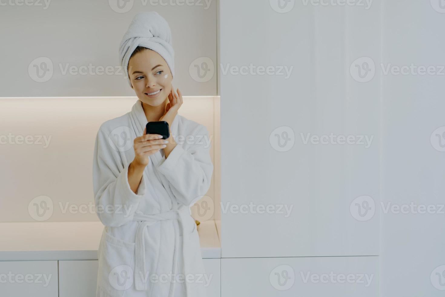 mulher atraente aplica creme hidratante no rosto cuida de sua tez e pele segura espelho usa roupão branco sorri gentilmente olha pensativamente posa sobre móveis modernos em casa foto