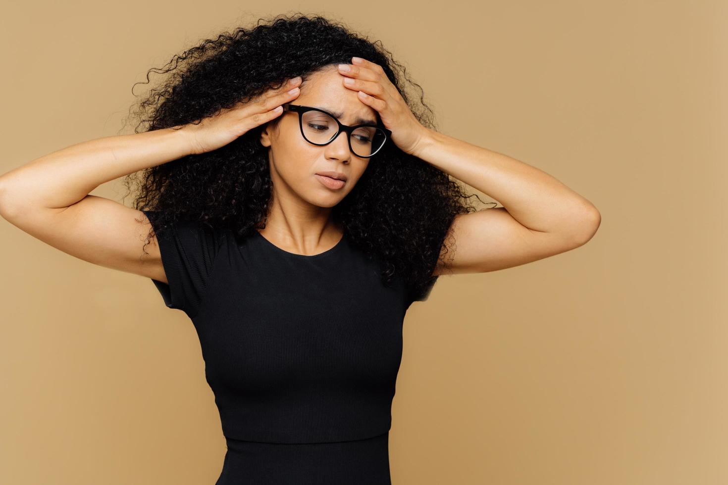 foto de meio comprimento de mulher estressante com penteado afro, focado para baixo, toca a cabeça, sofre de enxaqueca, cansado depois do trabalho, usa camiseta preta casual, focado para baixo, posa no estúdio marrom