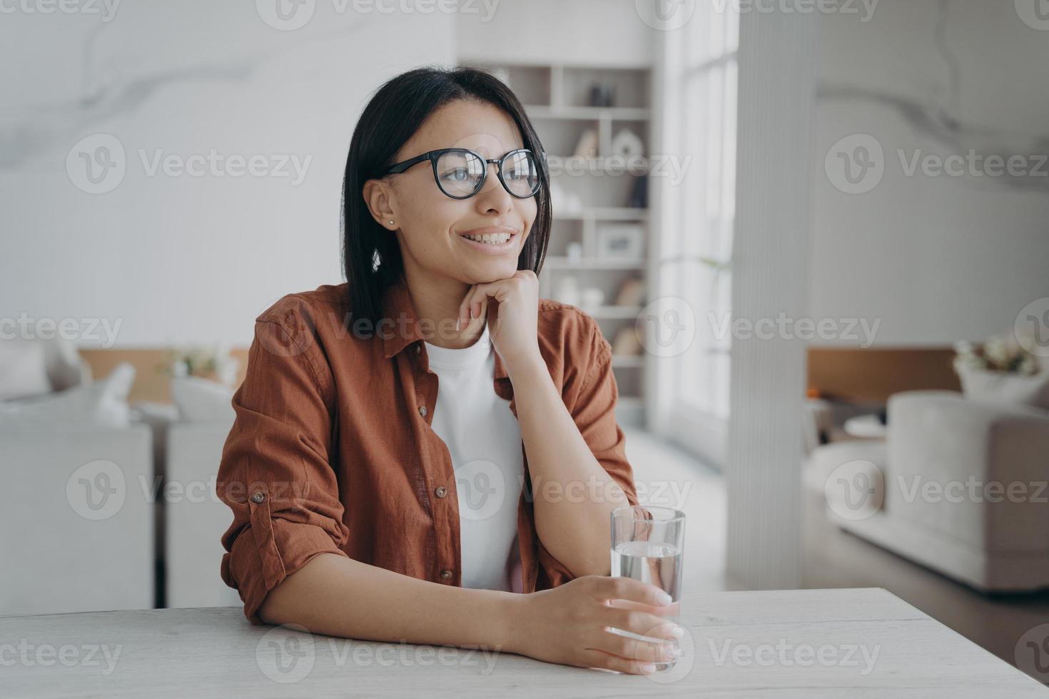 mulher sorridente desfrutar de água mineral purificada segurando o vidro sentado em casa. estilo de vida saudável, dieta foto