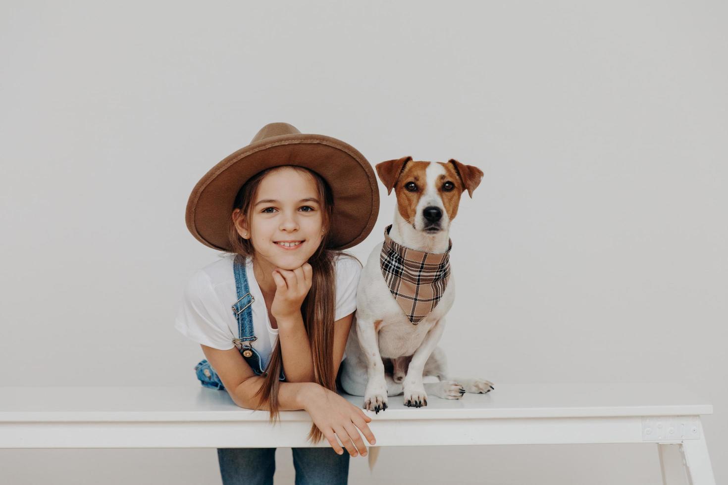 conteúdo menina bonita usa chapéu, se inclina na mesa branca, posa perto de cachorro com pedigree, gosta de passar o tempo livre juntos, vai passear, descansar em casa. crianças, felicidade, animais, conceito de estilo de vida foto