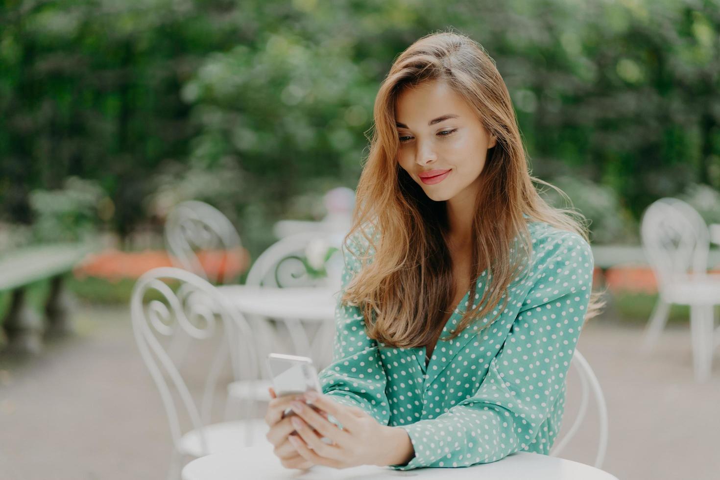 mulher bonita com maquiagem, vestida com roupas da moda, vestida com camisa de bolinhas da moda, usa o celular para comunicação online, navega nas redes sociais, posa na cafeteria ao ar livre foto