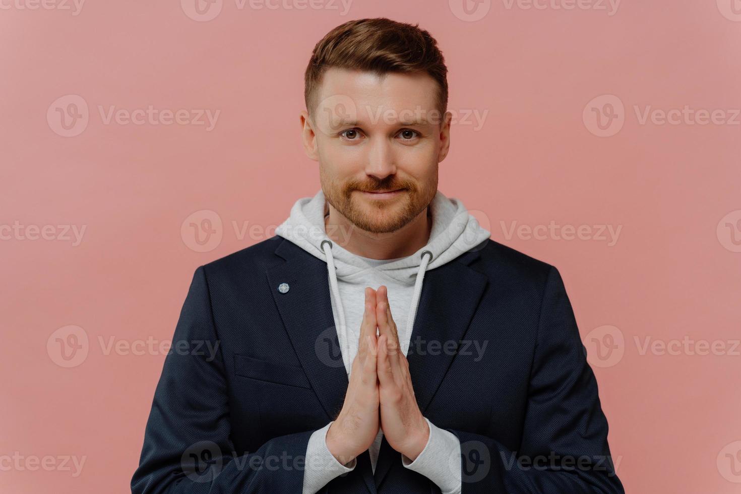 homem adulto barbudo sério mantém as palmas das mãos pressionadas juntas reza por algo olha diretamente para a câmera acredita em algo bom acontecer poses contra fundo rosa. cara bonito faz pedido foto