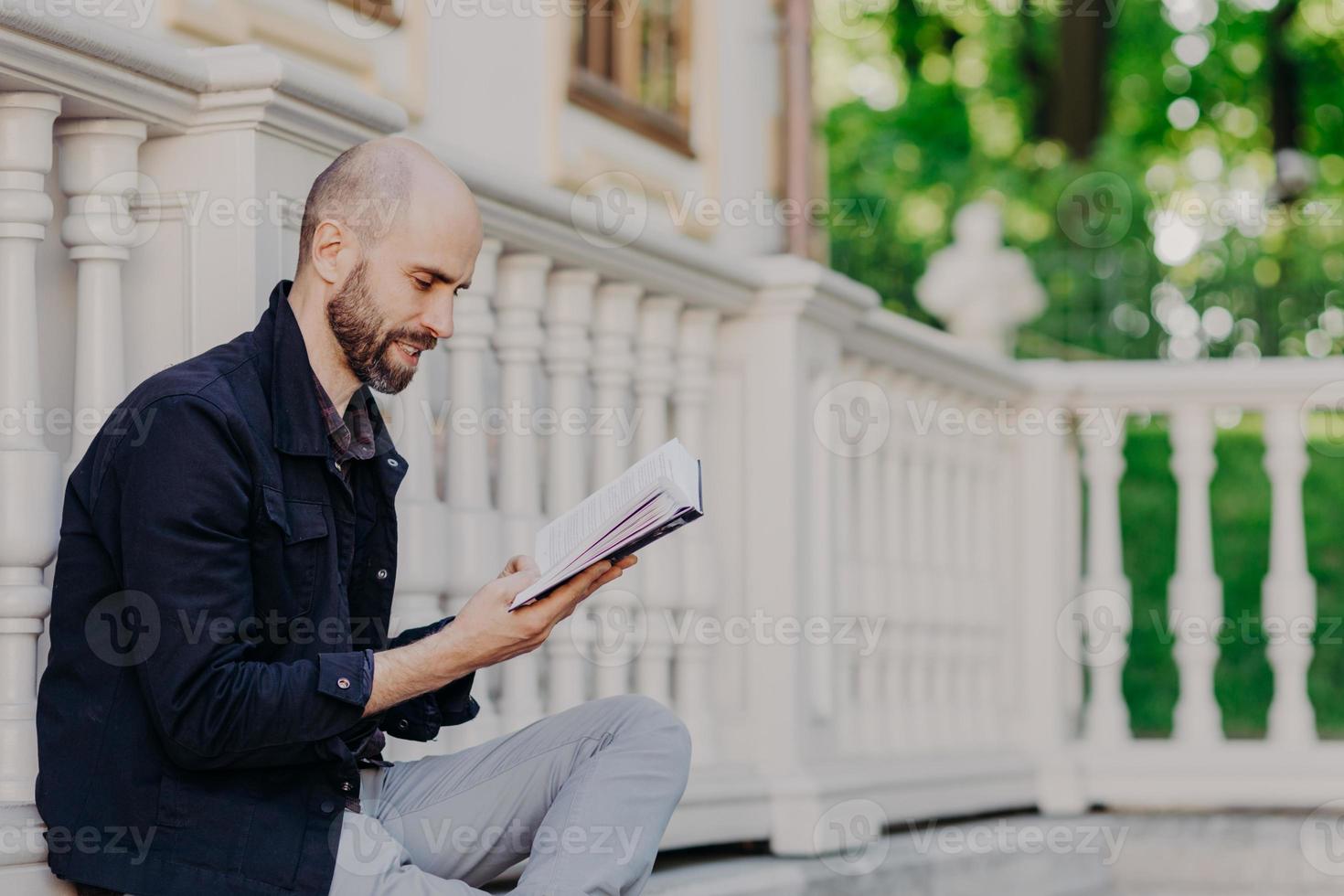 tiro lateral de homem barbudo com careca, segura o livro, senta-se ao ar livre contra o fundo da varanda branca, lê interessante história de detetive ou literatura científica. conceito de pessoas, educação e hobby foto