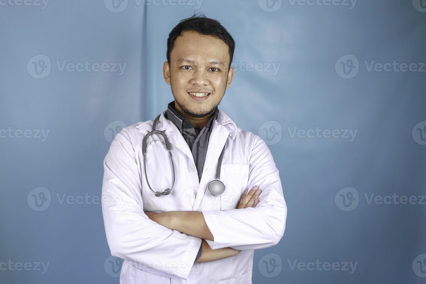 retrato de um médico masculino confiante atraente, braços sorridentes amigáveis cruzados usam estetoscópio de jaleco branco isolado fundo de cor azul foto