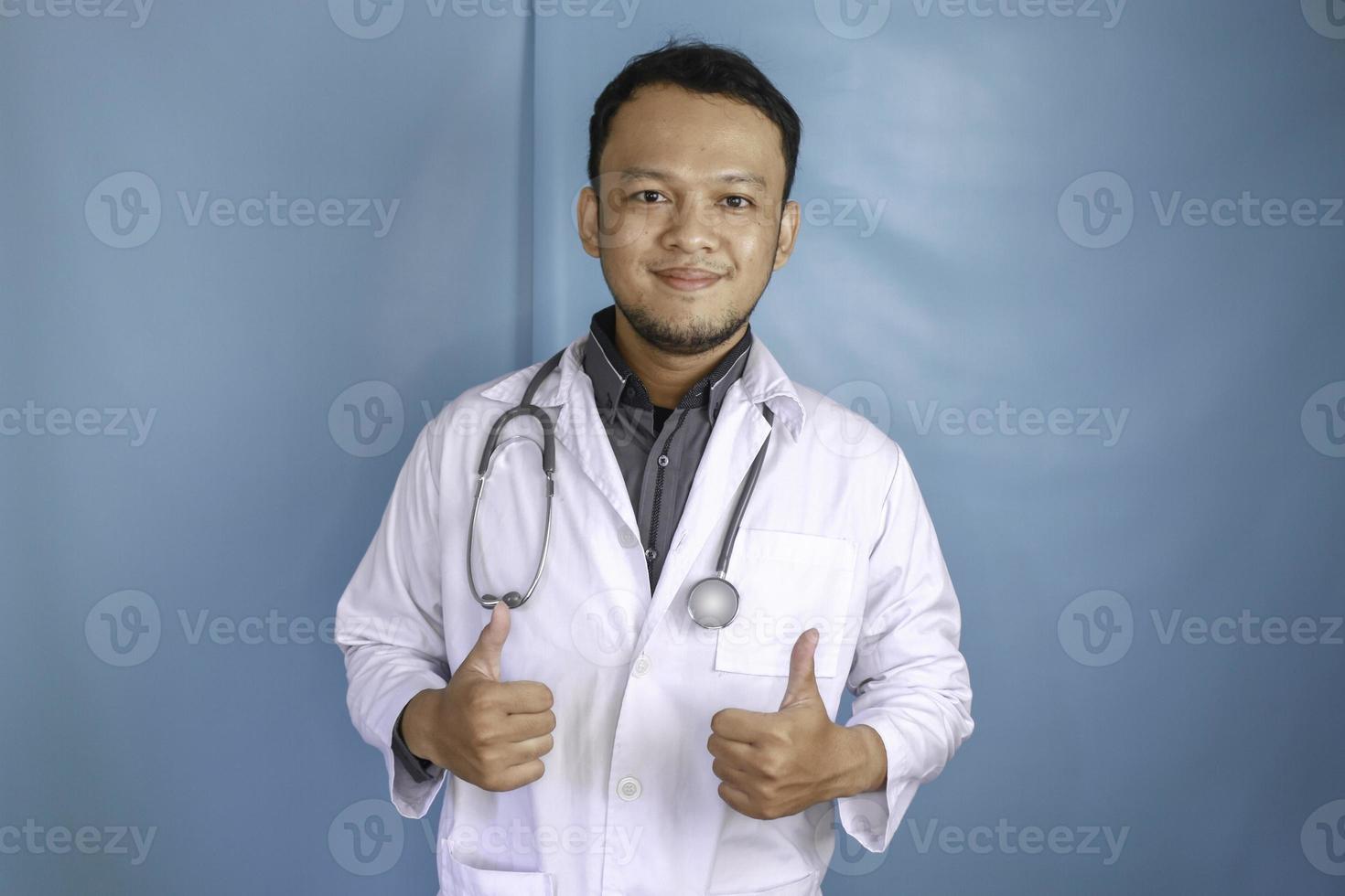 retrato de um médico jovem asiático, um profissional médico está sorrindo e mostrando os polegares para cima ou sinal de ok isolado sobre fundo azul foto