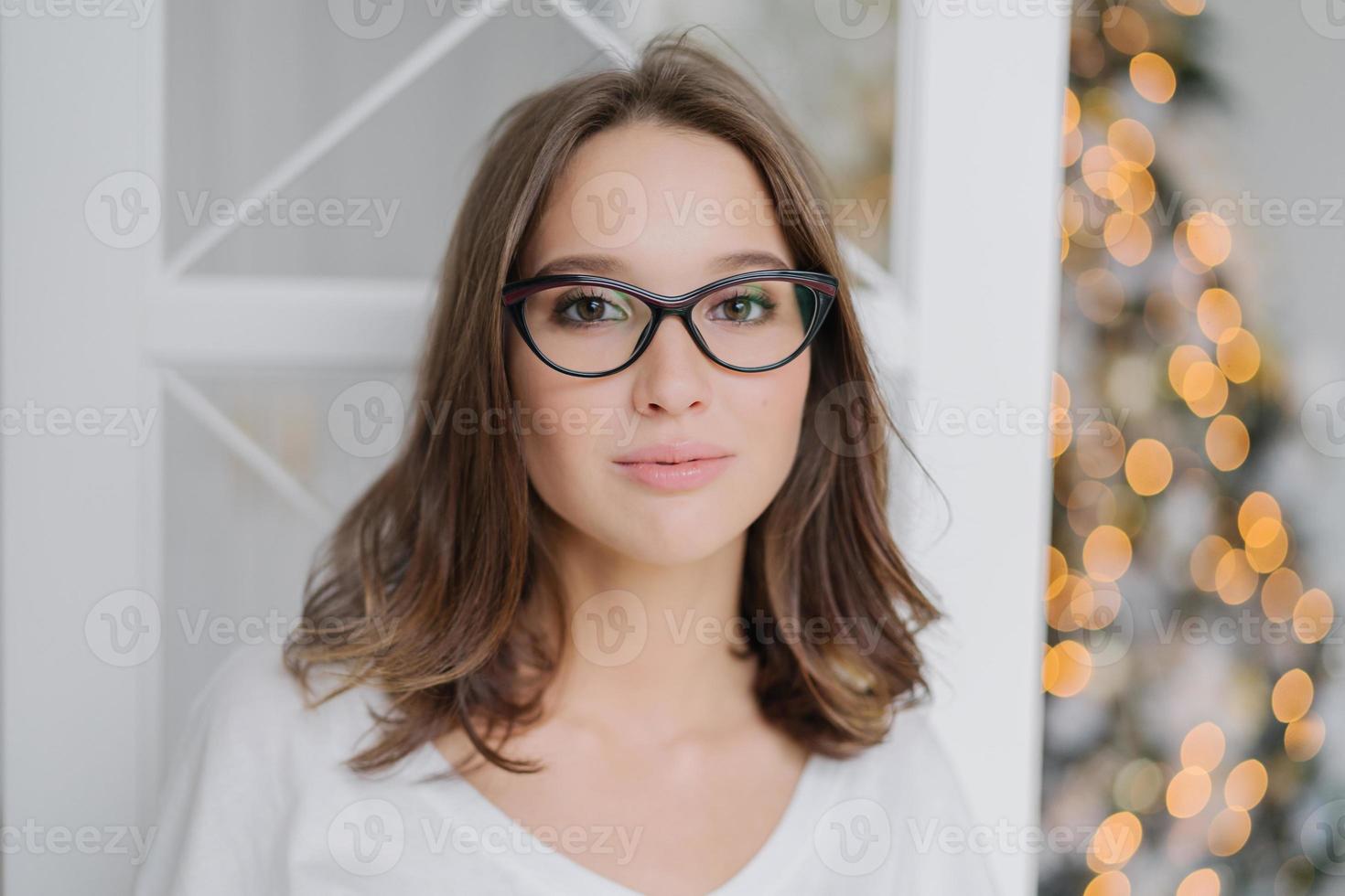 tiro na cabeça do adorável jovem modelo feminino em óculos transparentes, tem cabelos escuros, vestido com roupas casuais, gosta de atmosfera doméstica, posa contra a árvore do ano novo. conceito de pessoas e estilo de vida. foto