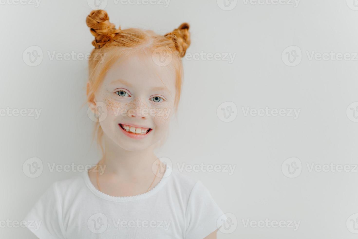 menina atraente positiva com pães duplos de gengibre, sardas no rosto, sorriso cheio de dentes, vestida com camiseta casual, fica contra um fundo branco, espaço vazio para o seu anúncio. conceito de crianças foto