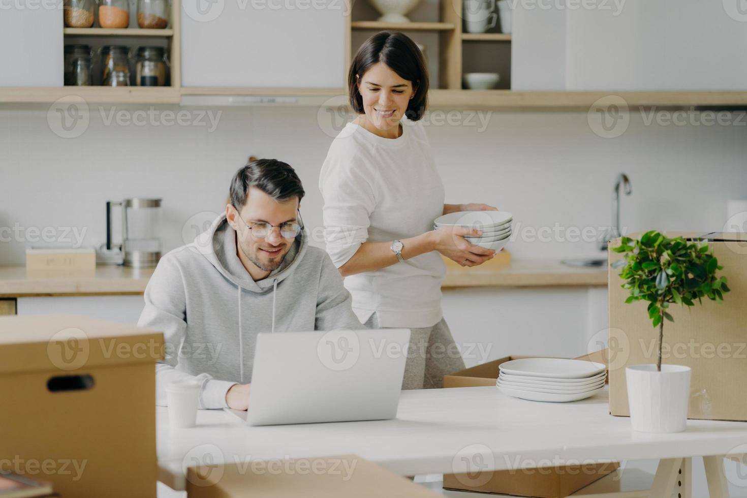 jovem morena desempacota caixas com pertences pessoais, coloca pratos no armário, olha atentamente para o computador portátil onde o marido trabalha. casal de família se muda para novo apartamento moderno foto