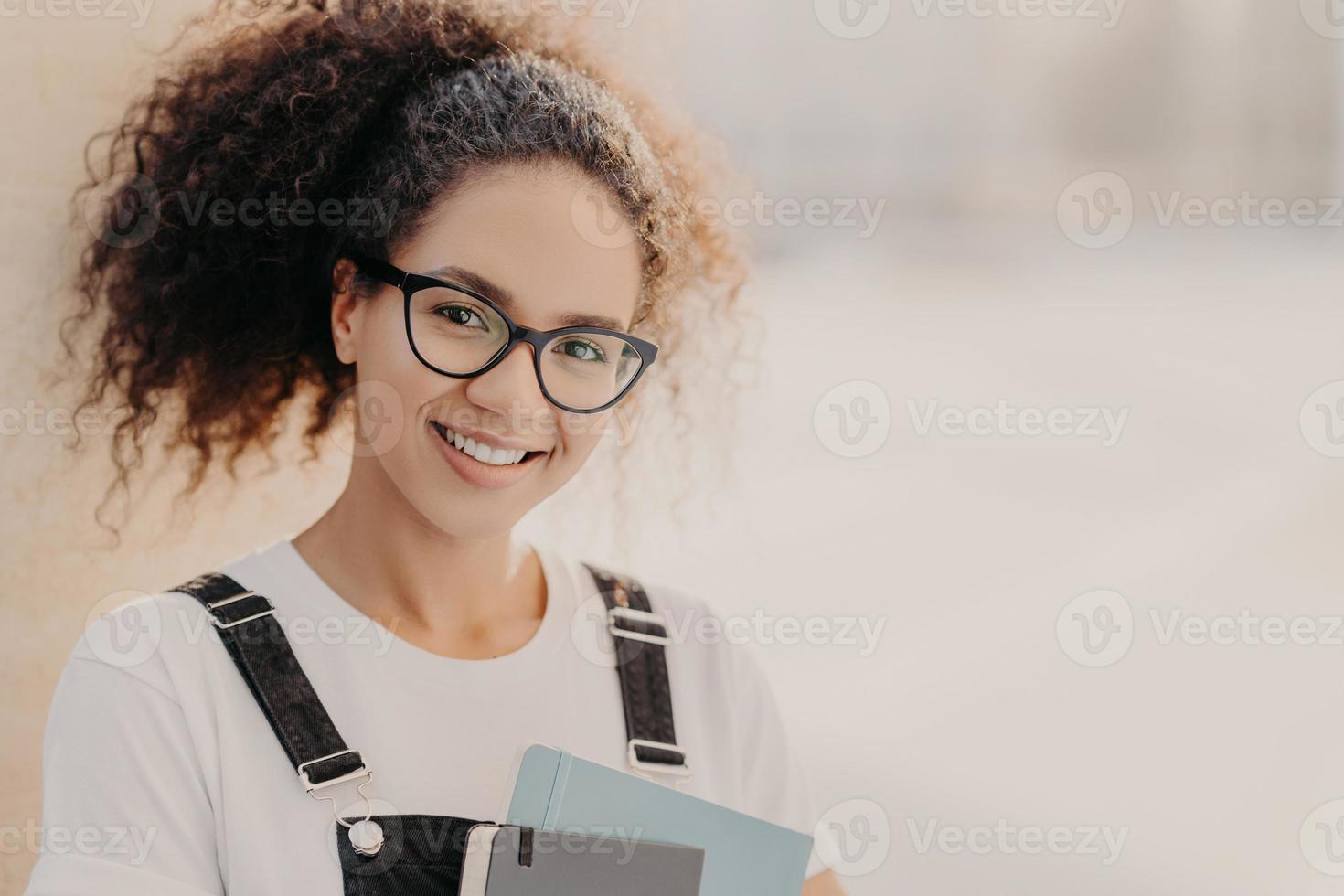 tiro horizontal de mulher bonita com cabelo crespo penteado em rabo de cavalo, carrega o bloco de notas, veste camiseta branca e macacão, gosta de estudar, posa contra um fundo desfocado com espaço vazio para texto foto