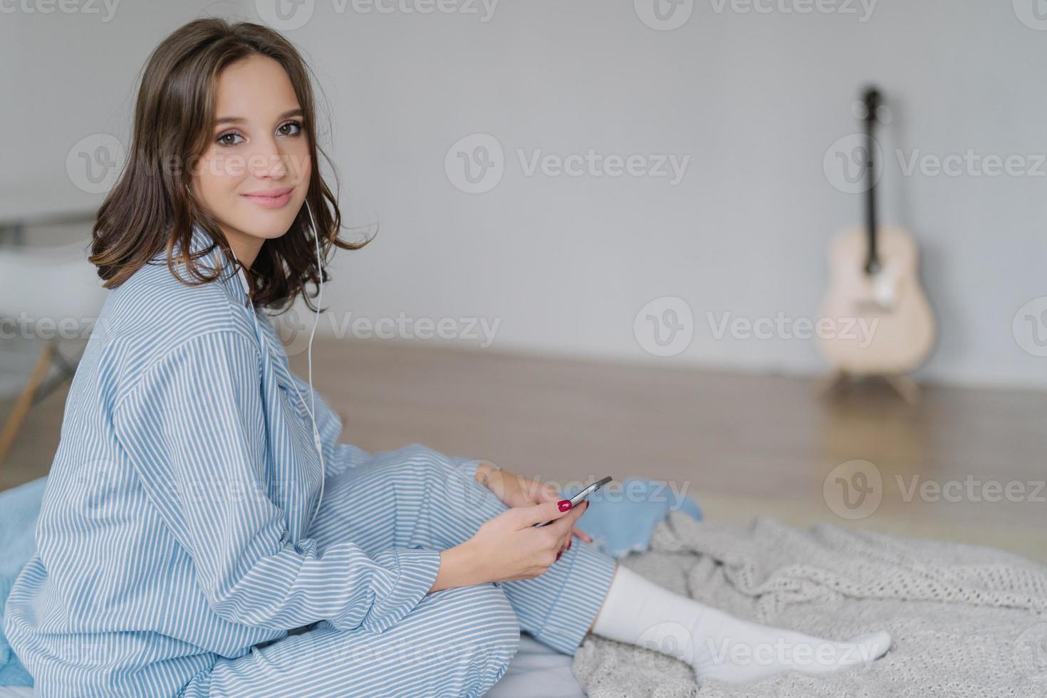 foto de mulher relaxada satisfeita, vestida com roupas casuais, senta-se na cama confortável, ouve música via aplicativo no smartphone e fones de ouvido, descansa no quarto sozinho, violão em segundo plano