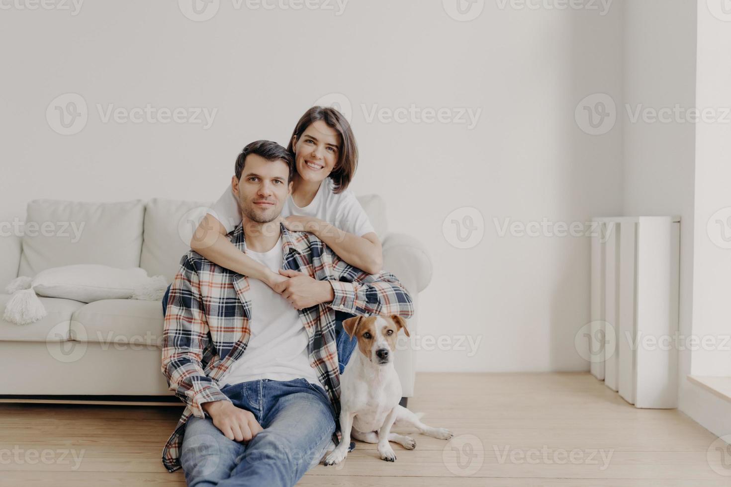 retrato de esposa afetuosa abraça marido que se senta no chão perto do sofá, seu animal de estimação favorito posa para a câmera, faz foto, estando em casa, compra apartamento novo, posa na sala de estar com paredes brancas foto