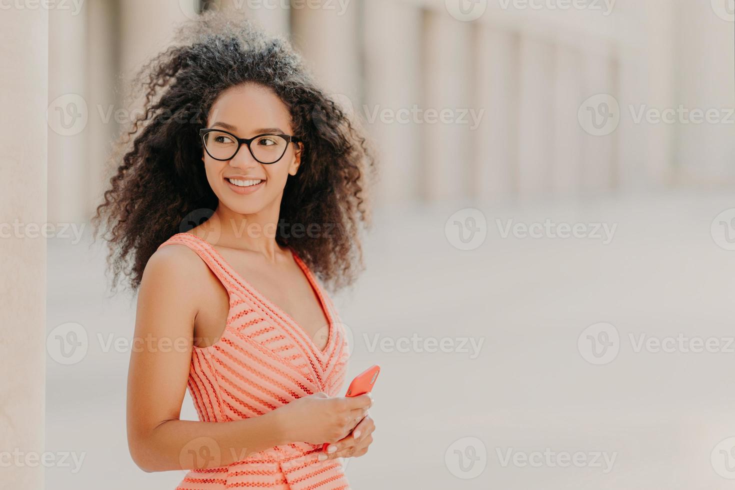 menina milenar satisfeita com cabelo encaracolado, usa vestido rosa, óculos ópticos, focado de lado com sorriso, fica ao ar livre, espera por ligação, gosta de conexão de internet de alta velocidade, atualiza novo aplicativo foto