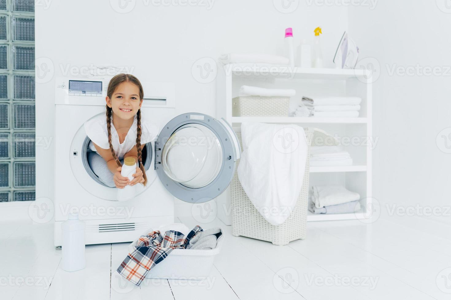 menina com aparência atraente, se diverte e posa dentro da máquina de lavar, segura detergente, se prepara para lavar, bacia com roupas para colocar na máquina de lavar no chão branco. dia de lavanderia em casa foto