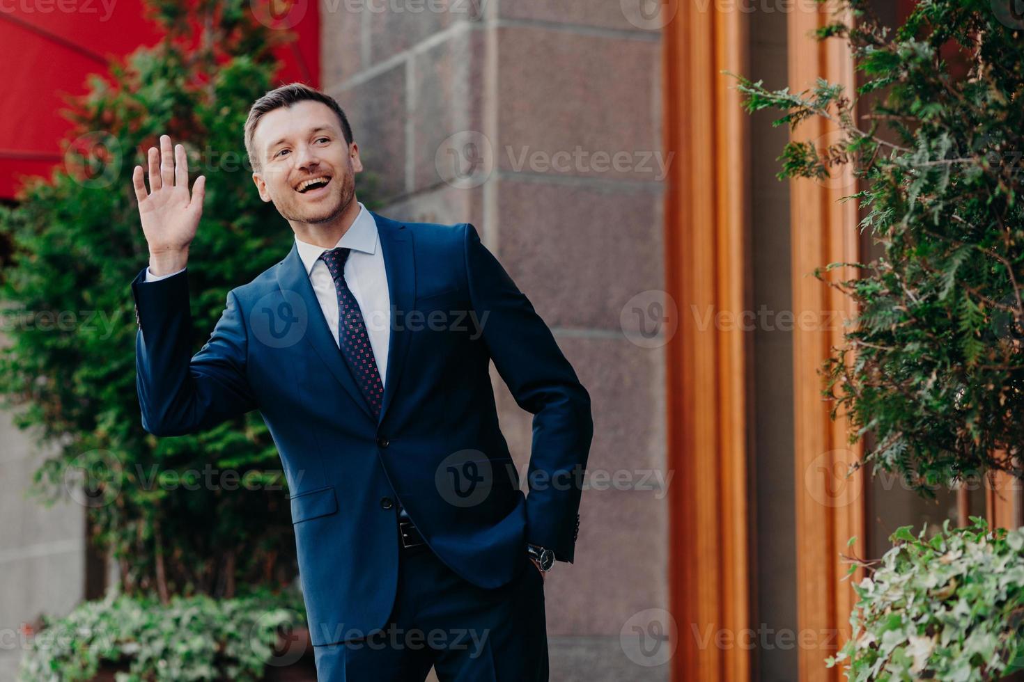 tiro de banqueiro masculino alegre em terno preto de luxo, acena com a mão como avisos amigo na rua, fica perto de seu escritório, vem trabalhar de bom humor, tem expressão positiva. pessoas, conceito de ocupação foto