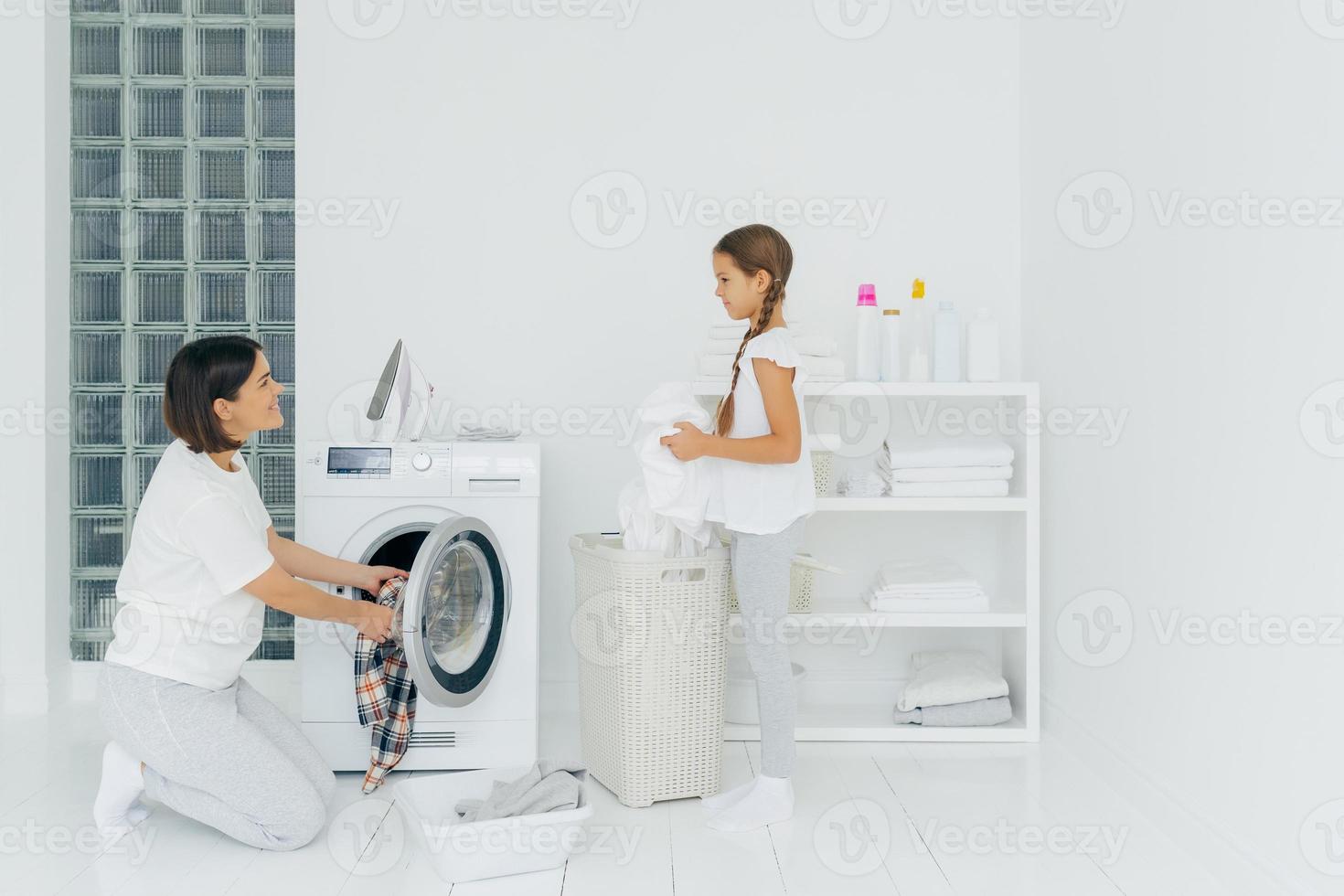 mulher morena ocupada carrega máquina de lavar com roupas sujas, sua filhinha ajuda, fica perto da cesta e classifica a roupa. feliz mãe e filho na sala de lavagem. conceito de trabalho doméstico. foto