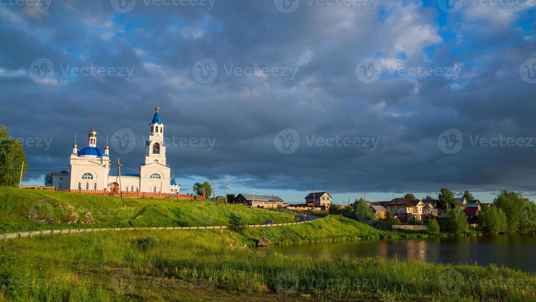 uma antiga igreja ortodoxa e uma vila na margem do rio. Sol e nuvens negras. foto