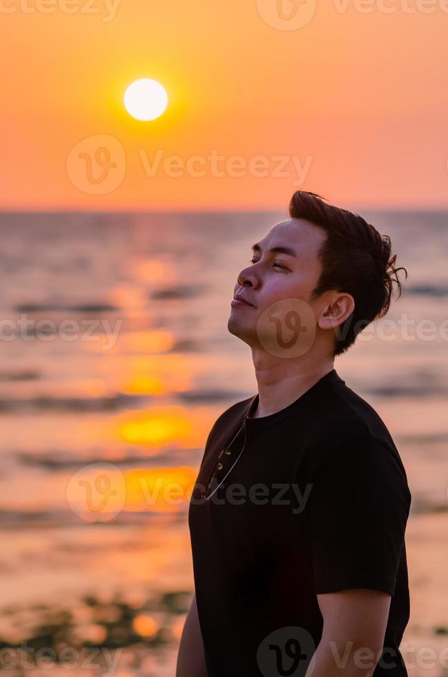 homem asiático olhando para o céu com o pôr do sol no fundo da praia. saúde mental positiva e expressão facial de emoção. foto