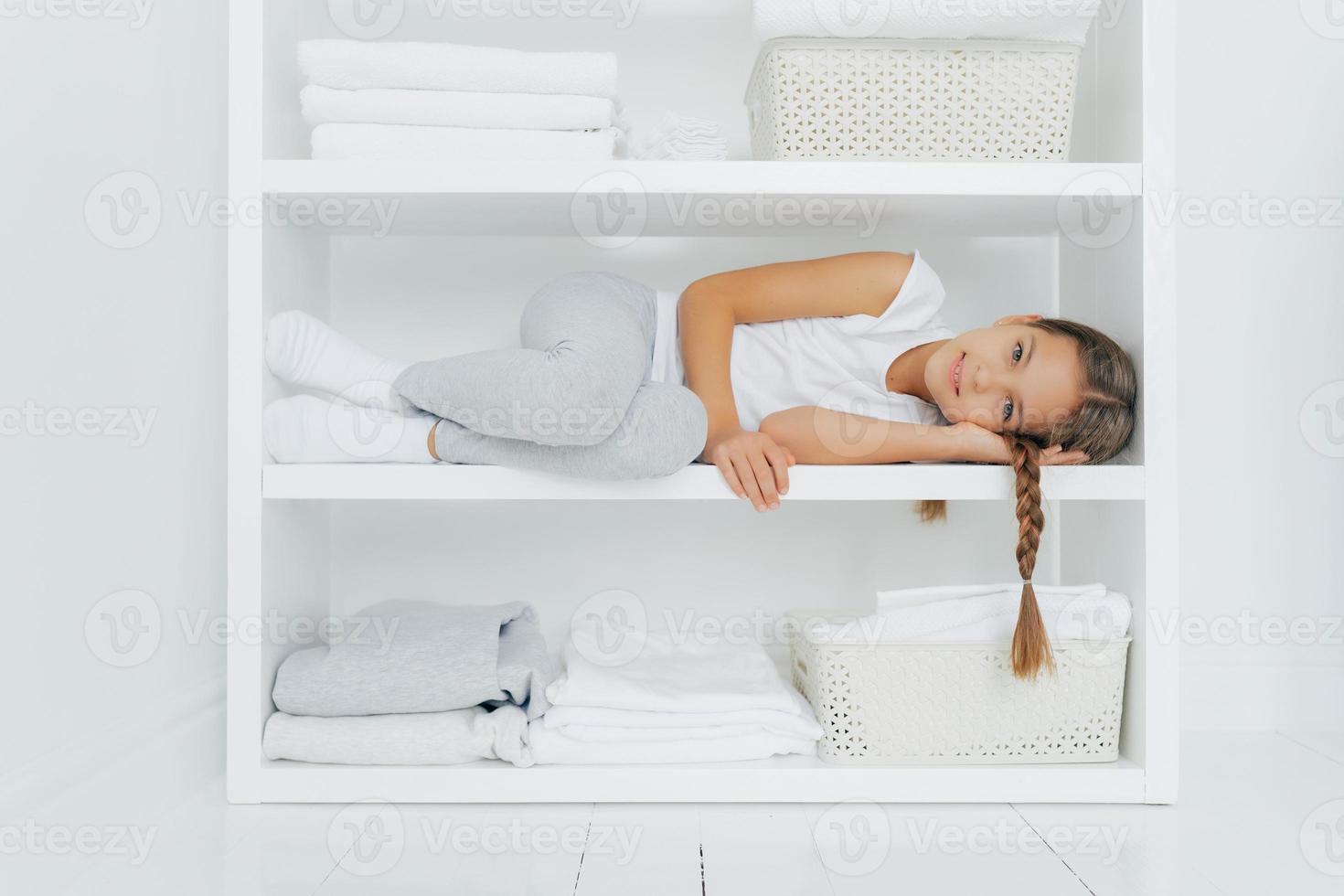 tiro horizontal de menina repousante com rabo de cavalo encontra-se na prateleira do console branco, vestido com roupas casuais, cercado com pilhas de linho dobrado, bacia de roupa fresca, tem descanso depois de ajudar a mãe foto