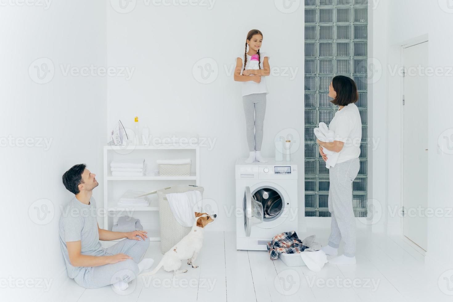 foto de marido e mulher, seu cachorro e filha fazem trabalhos domésticos na lavanderia, garotinha fica com detergente em cima da máquina de lavar, ocupada lavando roupas e lençóis, tem limpeza de primavera