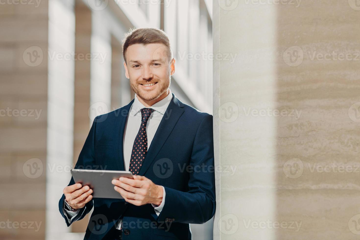 confiante alegre advogado masculino lê notícias de negócios, tem um sorriso gentil, vestido com roupas formais, posa em ambiente urbano. empresário verifica e-mail ou atualiza o perfil no computador tablet digital foto