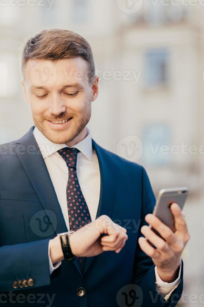 O proprietário de uma corporação masculina com barba por fazer feliz olha alegremente para o relógio de pulso, verifica a hora, recebe mensagem sms recebida no telefone inteligente, atualiza o perfil da conta bancária, usa conexão interent gratuita foto