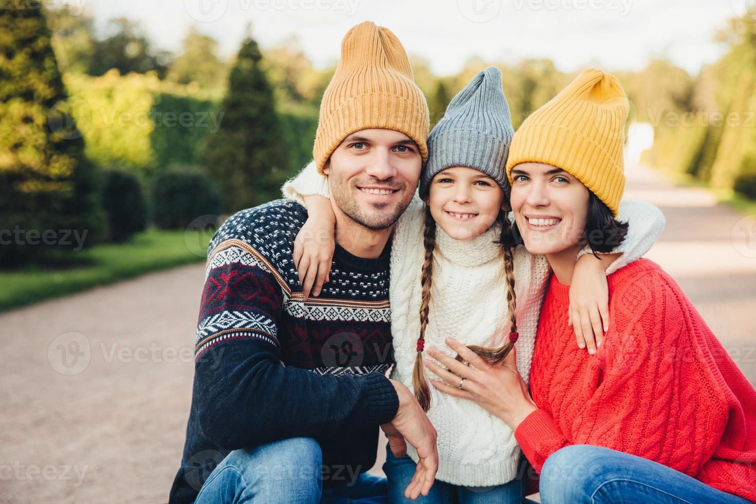 retrato horizontal de família afetuosa amigável se abraçam, usam bonés e suéteres de malha, caminham juntos, têm sorrisos agradáveis nos rostos. casal e filha pequena aproveitam o tempo livre foto